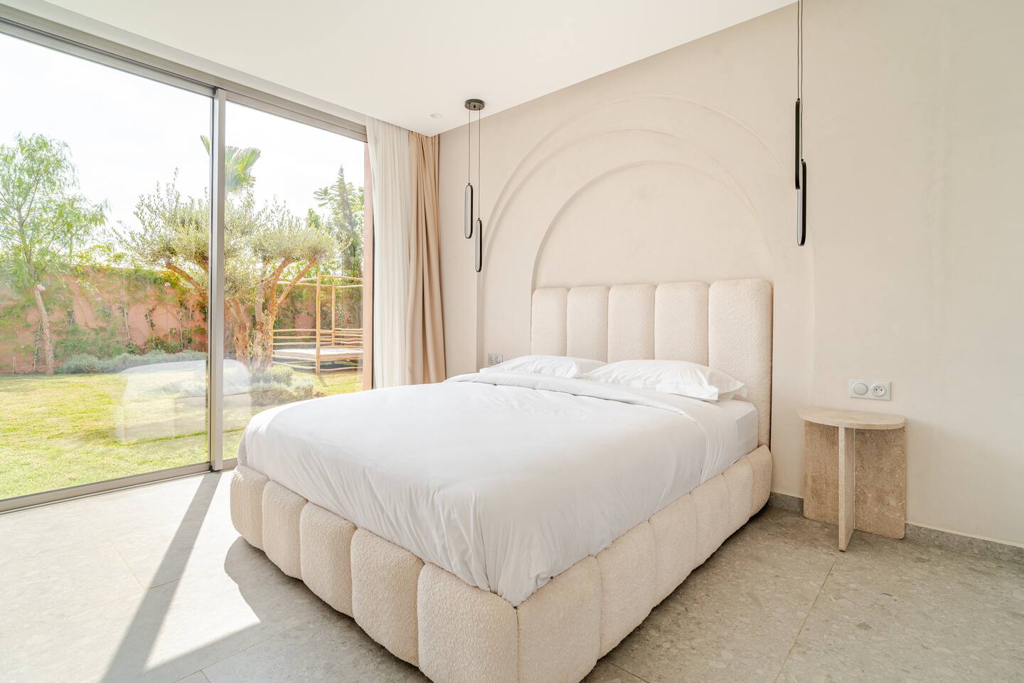 Bedroom with a cream upholstered king bed and a glass wall onto the garden
